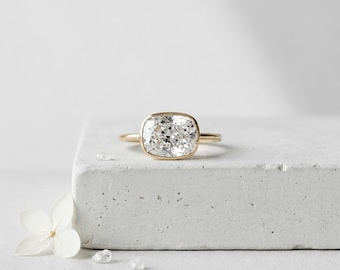 May include: A gold ring featuring a large, clear, cushion-cut gemstone set in a smooth bezel setting. The ring has a thin, polished gold band that encircles the finger. The gemstone displays multiple facets that reflect light, creating a brilliant appearance.
