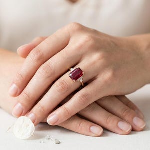May include: A gold ring featuring a large, rectangular, deep red gemstone set in the center, flanked by two smaller, clear, baguette-cut stones on each side. The band is thin and polished, showcasing a classic prong setting for the main stone.