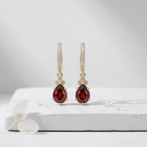 Può includere: Un paio di orecchini a cerchio color oro con un pendente a goccia di pietra preziosa rosso scuro. I cerchi sono incastonati con piccole pietre preziose trasparenti. Gli orecchini sono esposti su una superficie bianca.