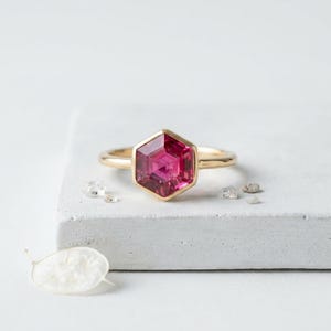 Könnte beinhalten: Ein goldener Ring mit einem sechseckigen, tiefrosa Edelstein. Der Ring ist in einem goldenen Band gefasst und liegt auf einer weißen Oberfläche. Kleine, klare Kristalle sind um den Ring herum verstreut.