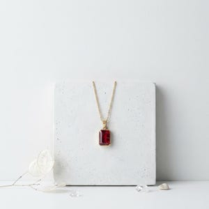 May include: A gold-colored necklace with a rectangular, deep red gemstone pendant. The pendant is set in a gold frame and hangs from a delicate gold chain. The necklace is displayed on a white surface.