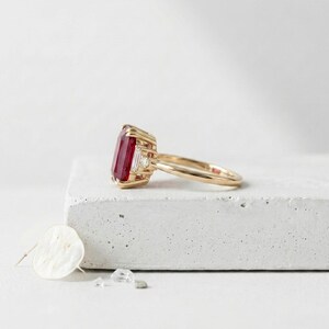 May include: A gold ring featuring a large, rectangular, deep red gemstone set in a prong setting. The ring includes two small, clear, baguette-cut accent stones on either side of the central red stone, mounted on a polished gold band.