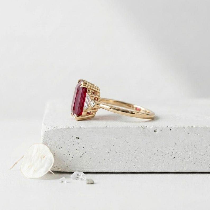 May include: A gold ring featuring a large, rectangular, deep red gemstone set in a prong setting. The ring includes two small, clear, baguette-cut accent stones on either side of the central red stone, mounted on a polished gold band.