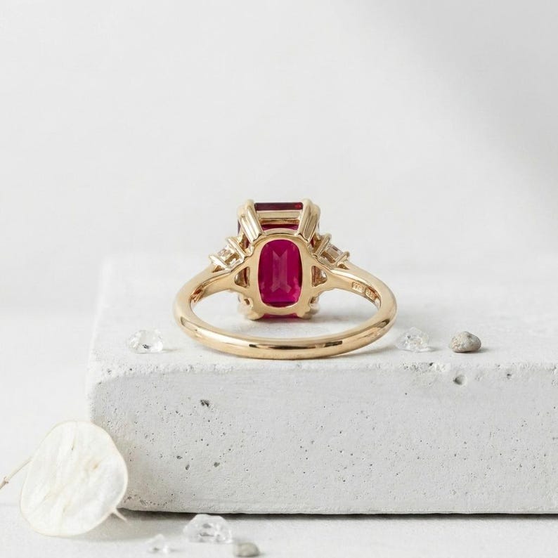 May include: A gold ring featuring a rectangular, emerald-cut vibrant pinkish-red gemstone set in a four-prong setting. The band is polished yellow gold, and the side of the ring displays intricate metalwork with small clear accent stones.