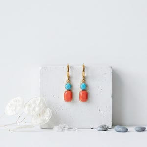 Puede incluir: Un par de pendientes de aro dorados con una pequeña cuenta redonda de color turquesa y una piedra rectangular de color coral. Los pendientes cuelgan de un aro de metal dorado, mostrando un contraste entre las piedras azules y naranjas.