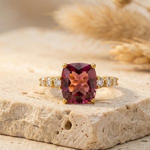 Puede incluir: Un anillo de oro con una gran piedra preciosa rosa rojiza de talla cuadrada. El anillo está adornado con piedras preciosas más pequeñas y claras a ambos lados de la piedra principal. El anillo está sobre una superficie clara y texturizada.
