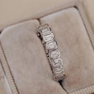 May include: A silver ring with multiple emerald-cut stones, set in a row. The ring is displayed in a gray velvet ring box. The background is a light beige color.