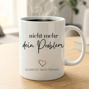 Könnte beinhalten: Weiße Keramik-Tasse gefüllt mit dunklem Kaffee, mit aufsteigendem Dampf. Die Tasse hat schwarzen Text, der „nicht mehr dein Problem“ und „SCHMECKT NACH FREIHEIT“ mit einem Herzsymbol lautet. Die Tasse steht auf einer Holzoberfläche.