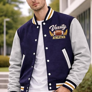 Puede incluir: Chaqueta universitaria azul marino y gris con detalles blancos. La chaqueta muestra el texto "Varsity Athletics" y un gráfico de fútbol americano. Tiene una botonadura y puños a rayas. La persona lleva una camiseta blanca y vaqueros azules.