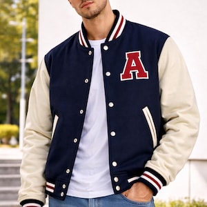 May include: Navy and cream varsity jacket featuring a large red "A" on the chest. The jacket has white snap buttons, white pockets, and striped cuffs. Worn over a white t-shirt and blue jeans.