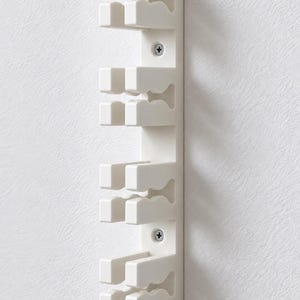 Peut inclure: Un organisateur mural vertical blanc doté de quatre fentes distinctes aux bords ondulés et profilés conçus pour maintenir des objets. L'unité est fixée au mur par deux vis métalliques argentées visibles situées entre les fentes.