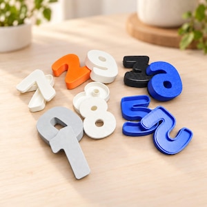 May include: A collection of three-dimensional plastic numbers in various colors including orange, white, black, blue, and silver. The set features individual digits such as 1, 2, 3, 5, 6, 7, 8, and 9, each with a flat base and raised numeral design.