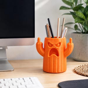 May include: An orange cylindrical pencil holder shaped like a cactus with a grumpy facial expression and two arms raised, each displaying a middle finger gesture. The holder contains several pens and pencils of various colors and metallic finishes.