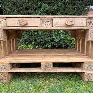 May include: A wooden pallet table with two drawers and a lower shelf. The table is constructed from reclaimed wood and features the "EPAL" stamp. The natural wood colour and simple design give it a rustic, industrial appearance. The table's dimensions are approximately 120 cm wide.