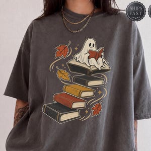 May include: A charcoal gray short-sleeve t-shirt featuring a graphic of a white ghost sitting on a stack of books and reading. The design includes colorful autumn leaves in shades of orange and yellow, small star accents, and a vintage-style wash.