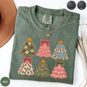 May include: A moss green crew neck t-shirt featuring a graphic print of six stylised Christmas trees. The trees are decorated with various patterns including polka dots, hearts, bows, and stars in shades of pink, blue, white, and green.