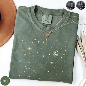 May include: A moss green t-shirt with a celestial design featuring gold stars, a crescent moon, and constellations. A silver necklace with a pendant rests on the shirt. The shirt is labeled "Comfort Colors" and is size S-P-CH.