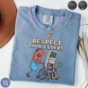 Könnte beinhalten: Ein blaues T-Shirt mit Rundhalsausschnitt und einer Grafik, die einen modernen Game-Controller zeigt, der eine alte Spielkonsole im Rollstuhl schiebt. Der Text darüber lautet RESPECT YOUR ELDERS. Das Shirt hat ein Comfort Colors Etikett.