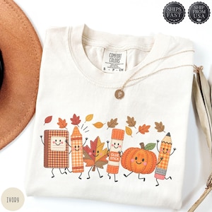 May include: An ivory t-shirt featuring a graphic print of anthropomorphic school supplies and a pumpkin with smiling faces, including a plaid notebook, a crayon, a maple leaf, a glue stick, a pumpkin, and a pencil, surrounded by falling autumn leaves.