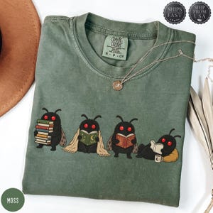 Peut inclure: Un t-shirt à col rond vert mousse orné d'un motif représentant quatre créatures noires aux yeux rouges semblables à des papillons de nuit lisant des livres. Une créature porte une pile de livres, une autre lit sous une couverture, une troisième tient un livre ouvert et la dernière est allongée avec un livre et une tasse.