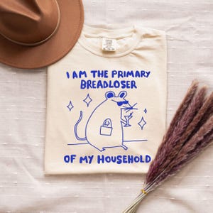 May include: Cream-colored t-shirt with the text "I AM THE PRIMARY BREADLOSER OF MY HOUSEHOLD" in blue. A cartoon rat wearing sunglasses and holding a cocktail glass is also printed on the shirt. A brown hat is visible in the upper left corner.