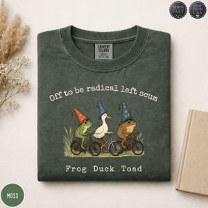 May include: A moss green t-shirt featuring a graphic of a frog, a duck, and a toad wearing wizard hats and riding bicycles. The text reads "Off to be radical left scum" and "FROG DUCK TOAD". The shirt has a Comfort Colors label at the inner neckline.