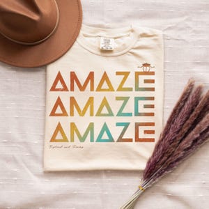 Könnte beinhalten: Cremefarbenes T-Shirt mit dem Wort "AMAZE" in einem sich wiederholenden Design aus Orange, Gelb und Türkis. Ein brauner Hut befindet sich in der oberen linken Ecke. Der Text "Ryland and Rocky" steht unten.