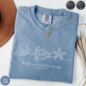 May include: A blue jean Comfort Colors t-shirt with a white seashell, turtle, and starfish design. The shirt features the text "Be still and know that I am God, Psalm 46:10". A gold necklace with a pendant is draped across the shirt.