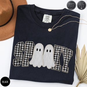 May include: A black short-sleeved t-shirt featuring the word SPOOKY appliqued in a black and white buffalo check pattern, with two white quilted ghost shapes replacing the letter O. The shirt has a crew neckline and a Comfort Colors brand label.