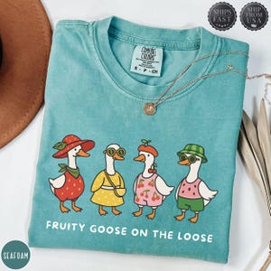 Könnte beinhalten: Türkisfarbenes T-Shirt mit einer Grafik von vier Gänsen in Outfits mit Obstmotiven. Die Gänse tragen einen Erdbeerhut, eine Sonnenbrille, ein Kirschbarett und einen Wassermelonen-Hut. Der Text "FRUITY GOOSE ON THE LOOSE" ist darunter gedruckt.