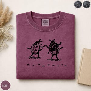 May include: Berry-coloured Comfort Colors crewneck sweatshirt with a black graphic of two stylised beetles holding hands, with ants below. The sweatshirt has a classic crew neck and a relaxed fit. The tag reads "Comfort Colors".