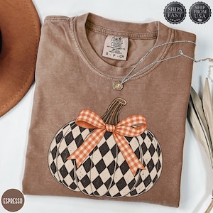 Könnte beinhalten: Ein espressofarbenes Kurzarm-T-Shirt mit einer Grafik eines Kürbisses mit schwarz-weißem Rautenmuster und einer orange-weiß karierten Schleife. Das T-Shirt hat einen Rundhalsausschnitt und ein Etikett der Marke Comfort Colors am Innenkragen.