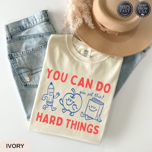 May include: An ivory t-shirt featuring the red text YOU CAN DO HARD THINGS and blue line art illustrations of a smiling pencil, apple, and book with the text you got this! The shirt is paired with light blue denim jeans and a tan wide-brim hat.