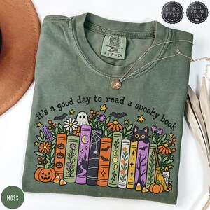 May include: A moss green crew neck t-shirt featuring a graphic print with the text 'it's a good day to read a spooky book'. The design includes a row of colourful illustrated books, a black cat, a ghost, pumpkins, and various floral elements.