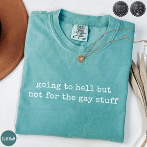 May include: Seafoam green t-shirt with the text "going to hell but not for the gay stuff" in white. The shirt has a crew neck and short sleeves. A gold necklace with a pendant is draped on the shirt.