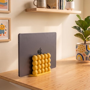 May include: A dark gray laptop held upright by a yellow stand featuring a grid of spherical, bubble-like protrusions. The stand is positioned on a light-colored wooden desk surface, providing a stable base for the closed electronic device.