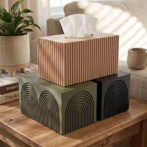 May include: A rectangular tissue box cover in a light tan color with vertical ribbed detailing, stacked atop two other rectangular boxes. One box is olive green with a repeating embossed arch pattern, and the other is solid black with the same arch design.