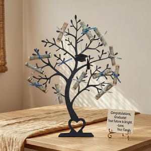 May include: A black metal graduation money tree with a heart-shaped base. Rolled-up dollar bills are attached to the branches with blue and silver ribbons. A small graduation cap hangs from the top. A card reads "Congratulations, Graduate!"