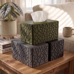 May include: Three decorative tissue box covers in shades of green and gray, with a textured, geometric pattern. One box is open with white tissues visible. The boxes are displayed on a wooden table, with a plant and mug in the background.