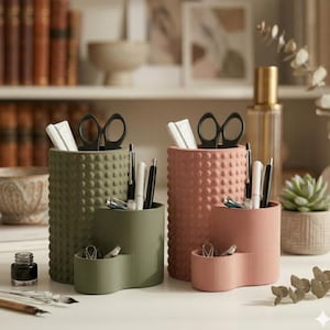 May include: Two desk organizers in olive green and dusty rose. Each has a textured, dotted surface and a two-tiered design. The organizers hold pens, scissors, and other office supplies. A small ink bottle and other stationery items are also visible.