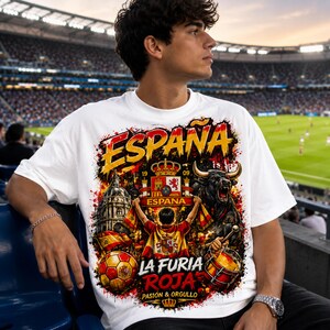 Può includere: T-shirt bianco con una grafica vivace. Il design include la parola "ESPANA", un toro, un pallone da calcio e il testo "LA FURIA ROJA". Il design include anche la bandiera spagnola e una corona.