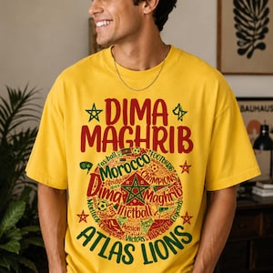 May include: A bright yellow short-sleeve t-shirt featuring a large circular graphic in red, green, and yellow. The text reads DIMA MAGHRIB at the top and ATLAS LIONS at the bottom, surrounding a central green star and various soccer-themed words.