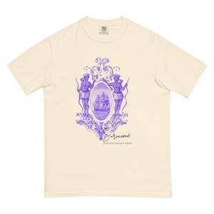 May include: Cream-colored t-shirt featuring a purple graphic. The graphic includes two figures with bows, a ship, and the word "Suriname" in cursive. The phrase "Justitia Pietas Fides" is below the word "Suriname".