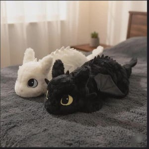 Könnte beinhalten: Zwei Plüschdrachen, einer weiß und einer schwarz, liegen auf einer grauen Oberfläche. Der schwarze Drache hat gelbe Augen und graue Flügel. Der weiße Drache hat blaue Augen. Beide haben weiches, strukturiertes Fell.