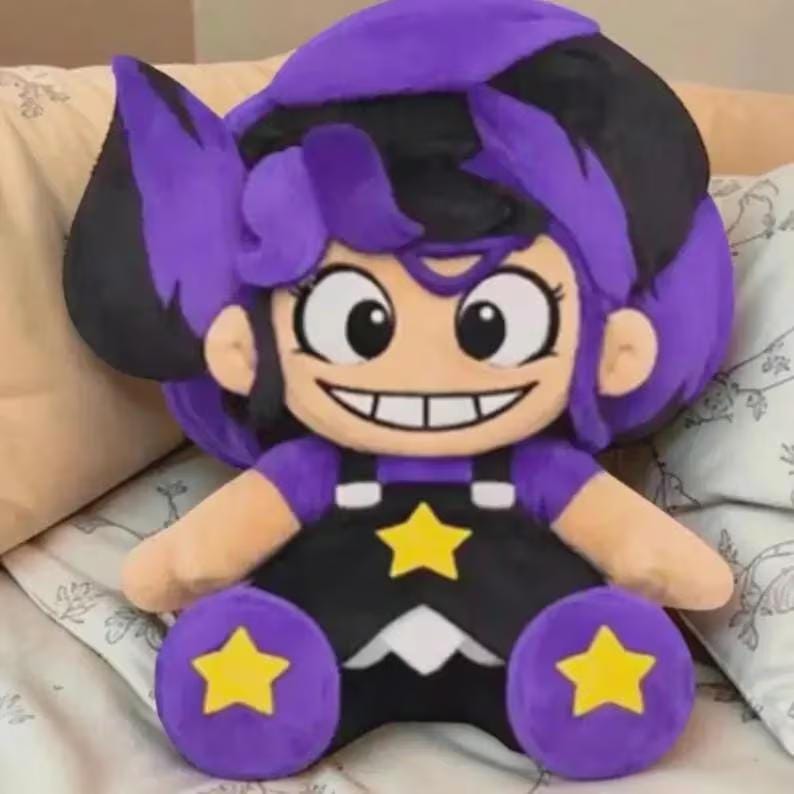 May include: A plush doll with a cartoon-style design. The doll has purple hair with black accents, large eyes, and a wide smile. It wears a black outfit with yellow star accents. The doll is sitting on a bed with a patterned pillow.