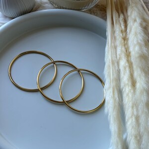 Może przedstawiać: Trzy złote bransoletki obręczowe ułożone na białym ceramicznym talerzu. Świeca zapachowa z napisem "Wood & Cashmere" i suszona trawa ozdobna w tle. Bransoletki mają prosty i elegancki design.