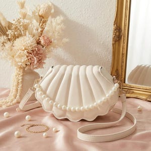 May include: A cream-coloured seashell-shaped handbag with a pearl-beaded trim and a long strap. The bag is made of a soft material and has a scalloped edge. A gold bracelet and pearls are scattered on a pink satin surface.