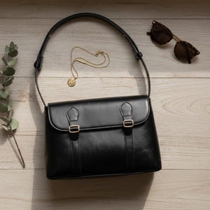 May include: A black leather satchel bag with gold-colored buckles and a long strap. A gold necklace with a pendant and tortoiseshell sunglasses are next to the bag on a wooden surface. A sprig of greenery is on the left.