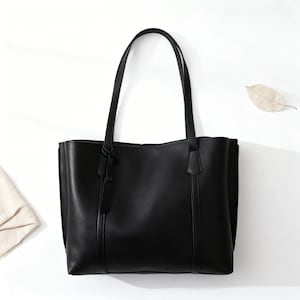 May include: A black tote bag with two long handles. The bag is made of a smooth material and has a rectangular shape. A small tag hangs from one of the handles. The bag is set against a white background.