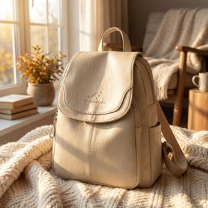 Puede incluir: Una mochila de color beige con cierre de solapa, asa superior y correas ajustables para los hombros. La solapa frontal presenta un logotipo de canguro en relieve y el texto Meishan Delshu. El bolso tiene un acabado liso y mate con bolsillos laterales.